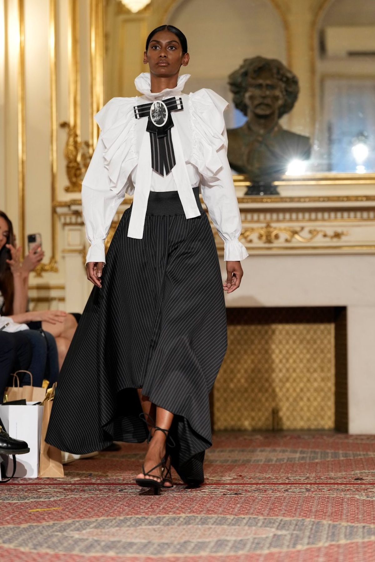 Модель у білій сорочці з рюшами та чорній спідниці під час показу Польського дня моди, стиль сучасної елегантності.