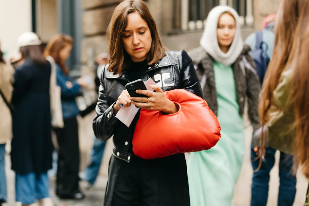 Продаж даних на Alibaba. Жінка з червоною сумочкою дивиться на свій телефон на жвавій міській вулиці. Ця сцена натякає на тему конфіденційності даних і на те, як часто ми ділимося інформацією в цифровому просторі.