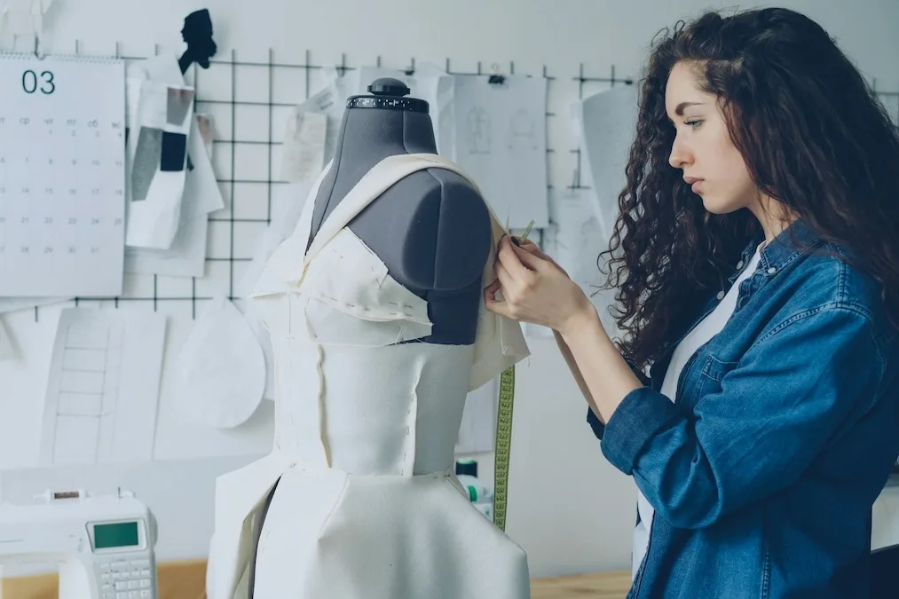 Дизайнерка приміряє сукню на манекені в кравецькому ательє під час створення колекції. Фахівці з практичними навичками конструювання та шиття користуються дедалі більшим попитом, що поступово призводить до підвищення їхніх зарплат.