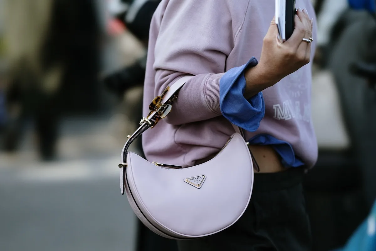 Аутфітерка вуличного одягу тримає на плечі світло-фіолетову сумку Prada, сфотографовану під час заходу вуличної моди. Де продавати одяг класу люкс онлайн?