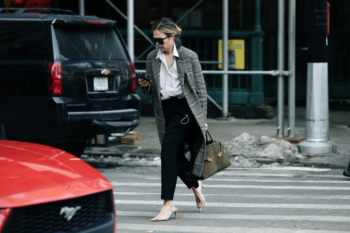 Жінка користується смартфоном, переходячи вулицю під час перевірки права L4 – ситуація, яку може розслідувати Установа соціального страхування (ZUS) у контексті оплачуваної роботи. У 2026 році перевірки лікарняних будуть детальнішими, і будь-яка діяльність може вплинути на право на отримання допомоги по хворобі.