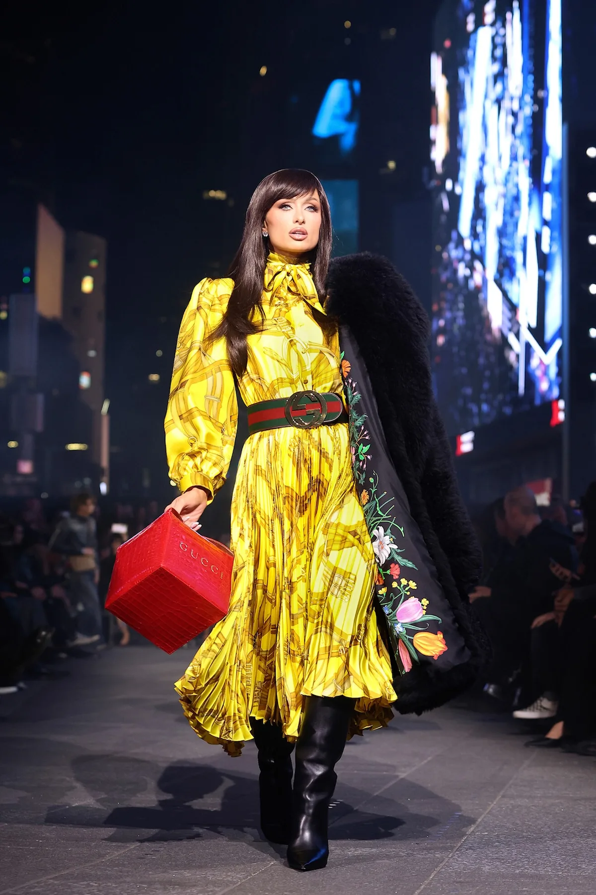 Періс Хілтон демонструє свій образ Gucci Resort S27 на нічному подіумі в центрі міста — жовту плісовану шовкову сукню, пояс з логотипом Gucci, чорні чоботи та червону косметичку.