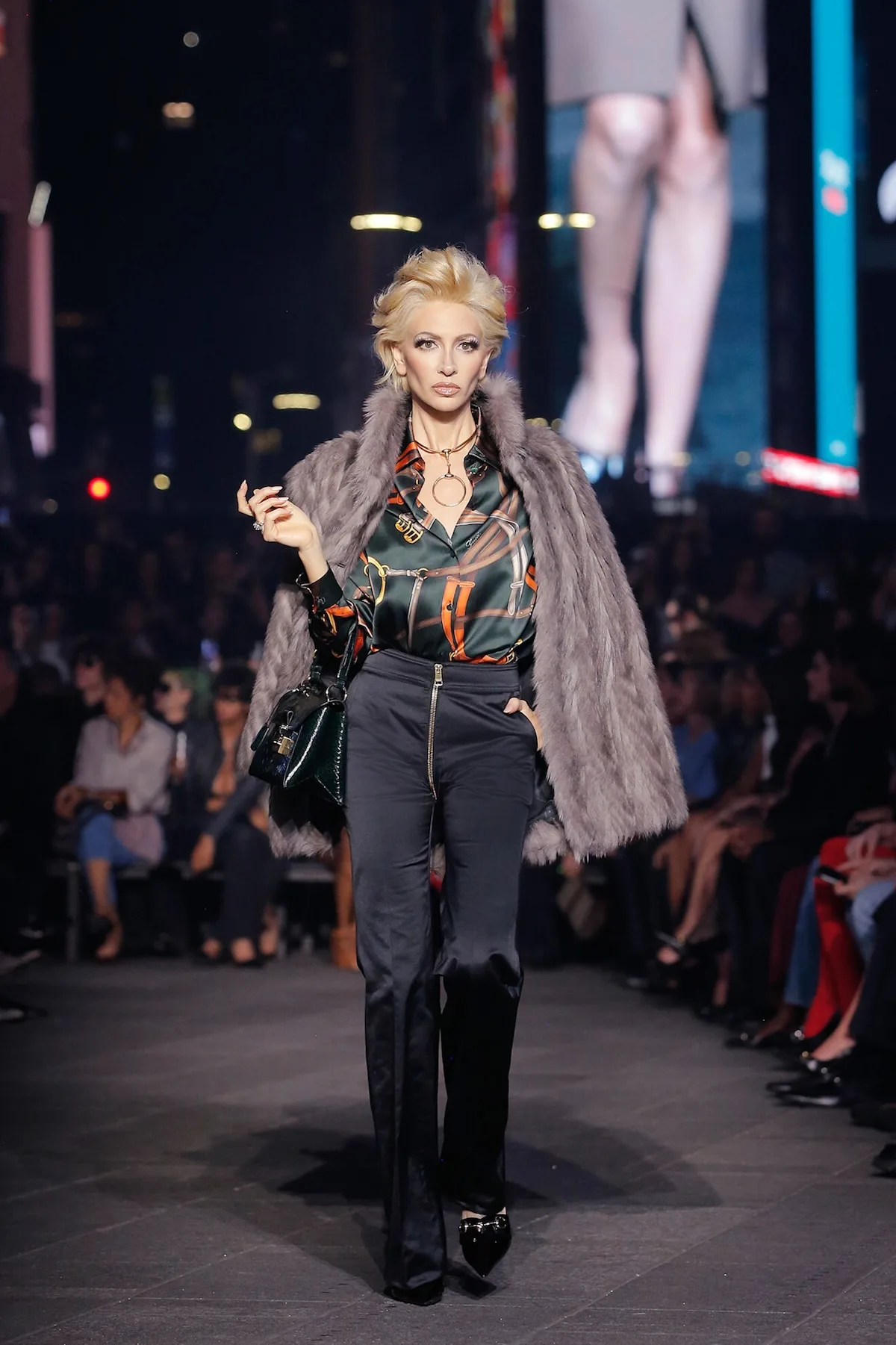 Модель на показі Gucci Resort S27 в елегантному вбранні з візерунчастою атласною сорочкою, чорними штанами з високою талією та сірим хутряним пальто, накинутим на плечі.